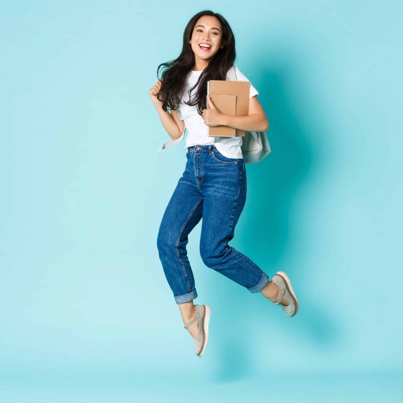 Fashion, back to school and lifestyle concept. Cheerful young asian girl, korean student looking upbeat, holding backpack and notebooks, jumping upbeat over light blue background